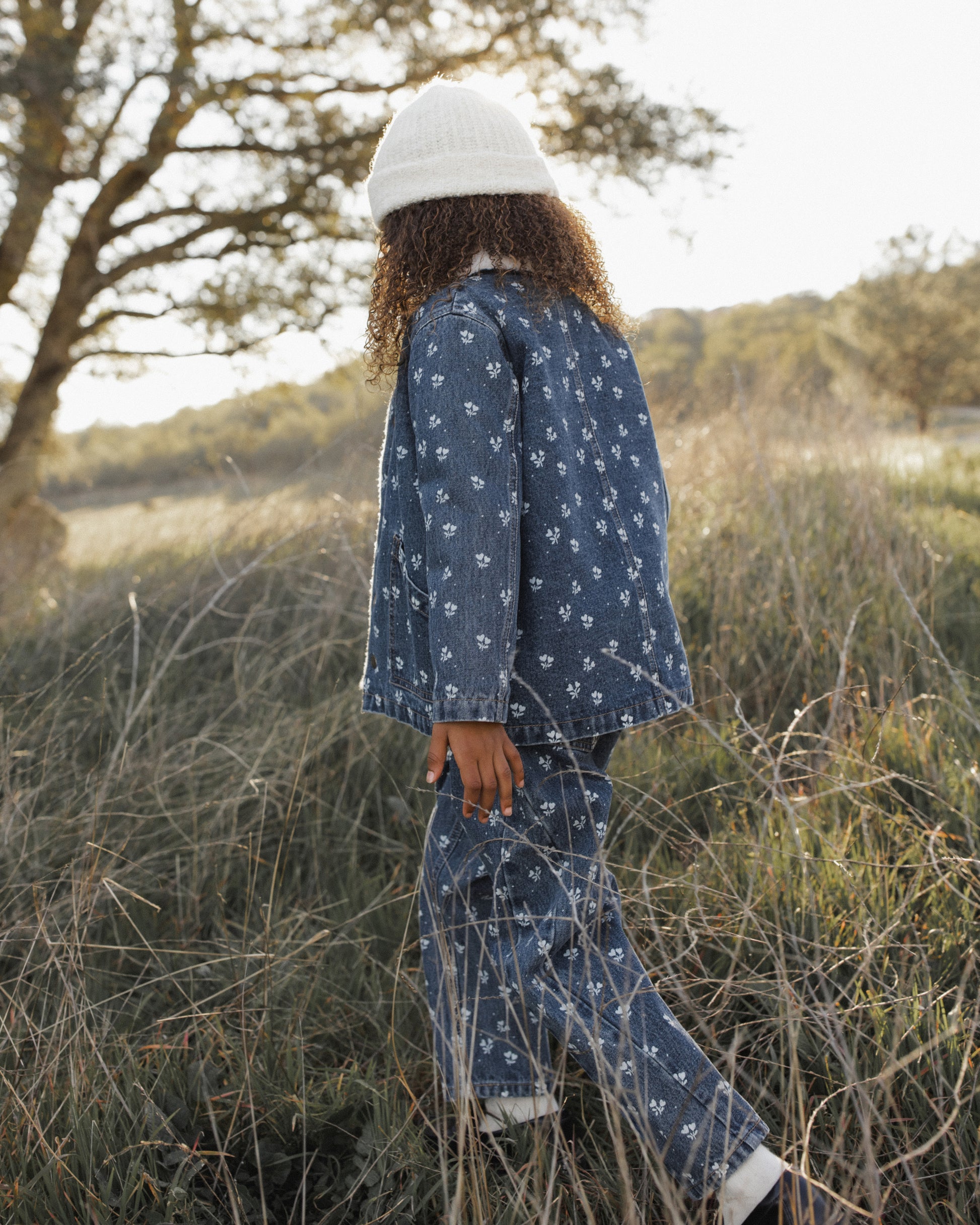 A child wearing a matching blue denim set with a small white floral print and a white knit beanie, standing in tall dry grass outdoors near trees.