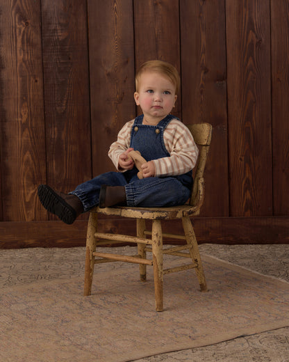 A toddler sitting on a rustic wooden chair in front of a dark wood plank wall, wearing blue denim overalls over a beige and brown striped long-sleeve shirt, with dark boots. The child is holding a small wooden toy.