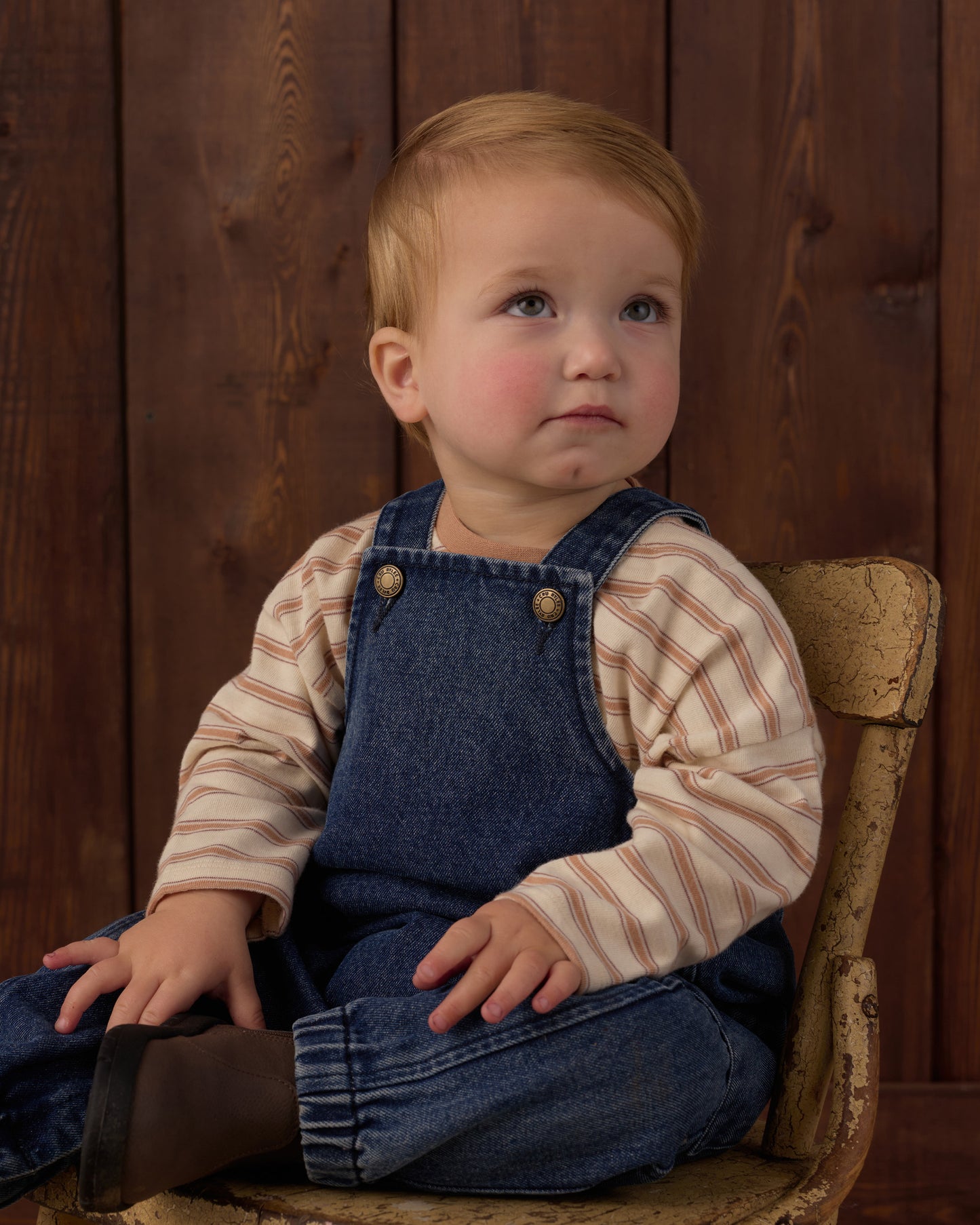 A baby boy with light hair sits on a rustic wooden chair against a dark wood plank background, wearing a beige and brown striped long-sleeve shirt under blue denim overalls.