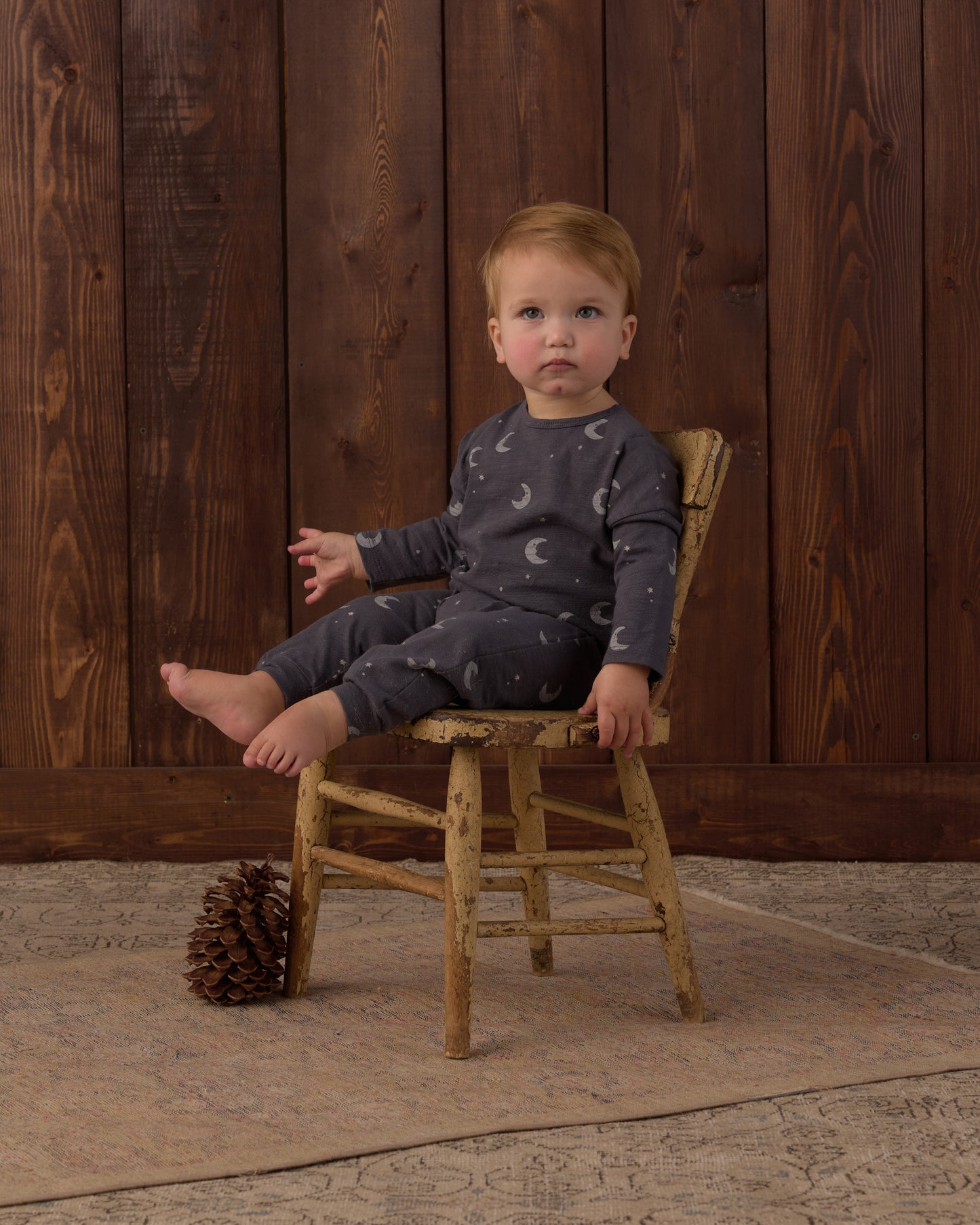 A toddler sits on a rustic wooden chair in front of a dark wood plank wall, wearing a dark gray long-sleeve pajama set with a white crescent moon pattern. A pinecone rests on a beige rug on the floor.
