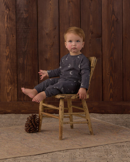 A toddler sits on a rustic wooden chair in front of a dark wood plank wall, wearing a dark gray long-sleeve pajama set with a white crescent moon pattern. A pinecone rests on a beige rug on the floor.