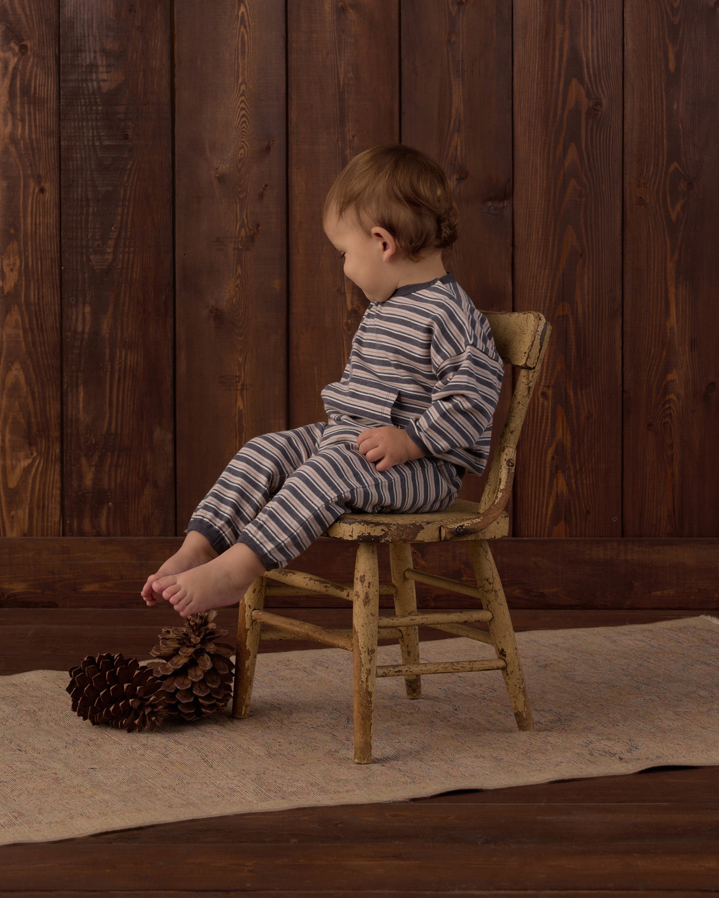A toddler wearing a matching striped long-sleeve top and pants set sits on a small rustic wooden chair in front of a dark wood plank wall. The outfit has thin dark stripes on a light gray or off-white base.