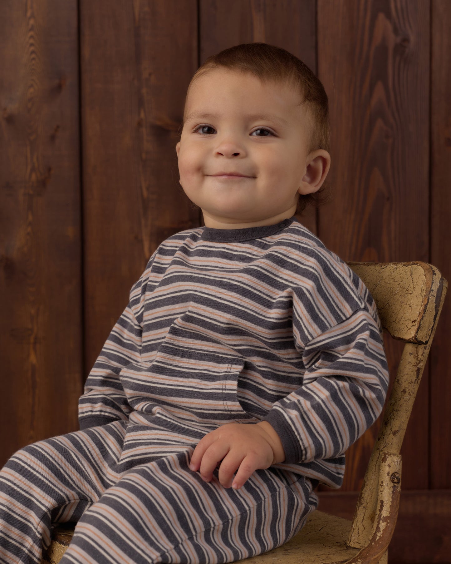 A smiling toddler boy wearing a navy and beige striped long-sleeve pajama set, sitting on a rustic wooden chair against a dark wood-paneled background.