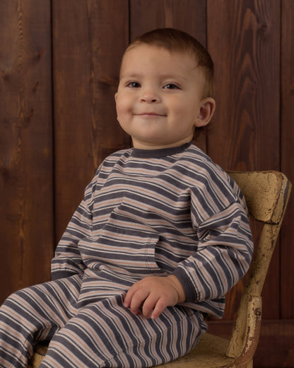 A smiling toddler boy wearing a navy and beige striped long-sleeve pajama set, sitting on a rustic wooden chair against a dark wood-paneled background.