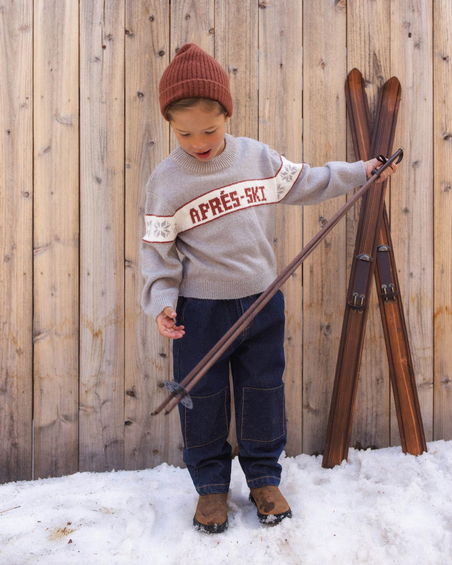 Child holding a pair of wooden skis in the snow, wearing the Reagan Apres Ski sweater and a rust beanie