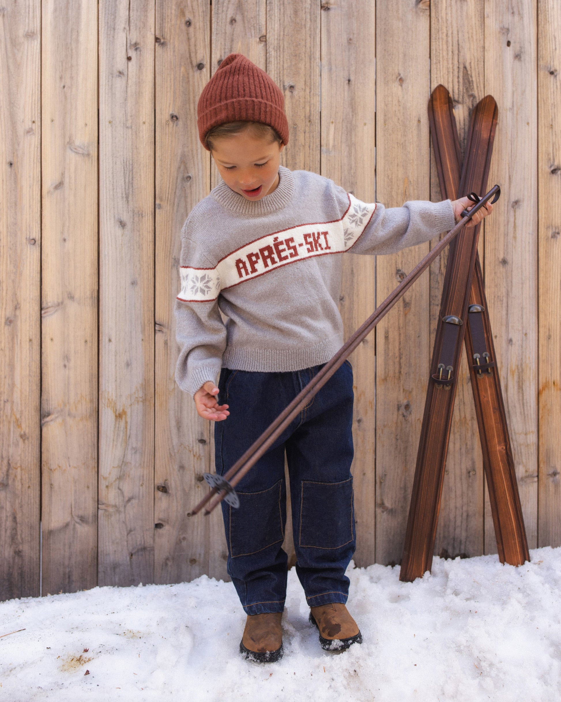 Child holding a pair of wooden skis in the snow, wearing the Reagan Apres Ski sweater and a rust beanie