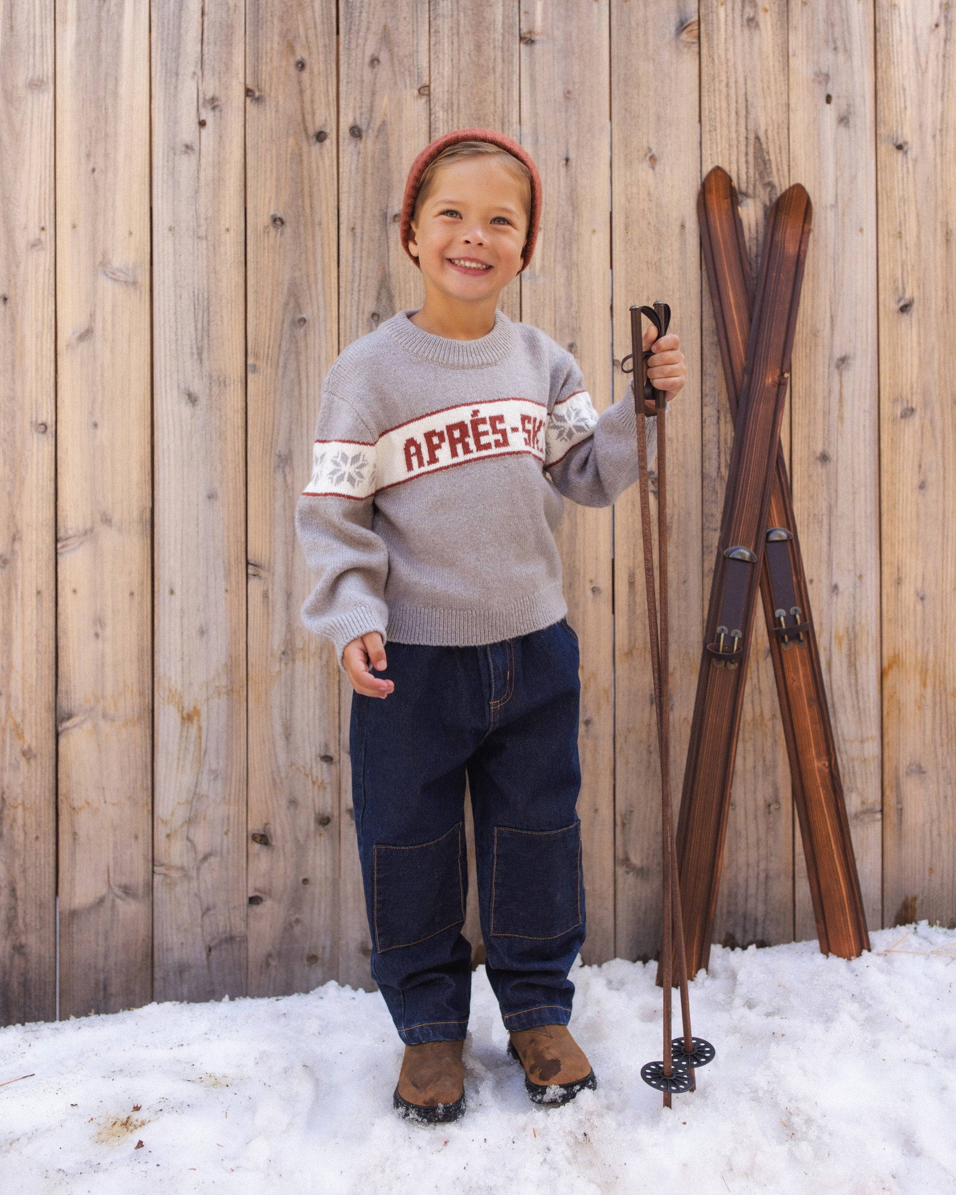 Child in a rust beanie standing in the snow beside vintage wooden skis, wearing the Rylee + Cru Reagan Apres Ski sweater