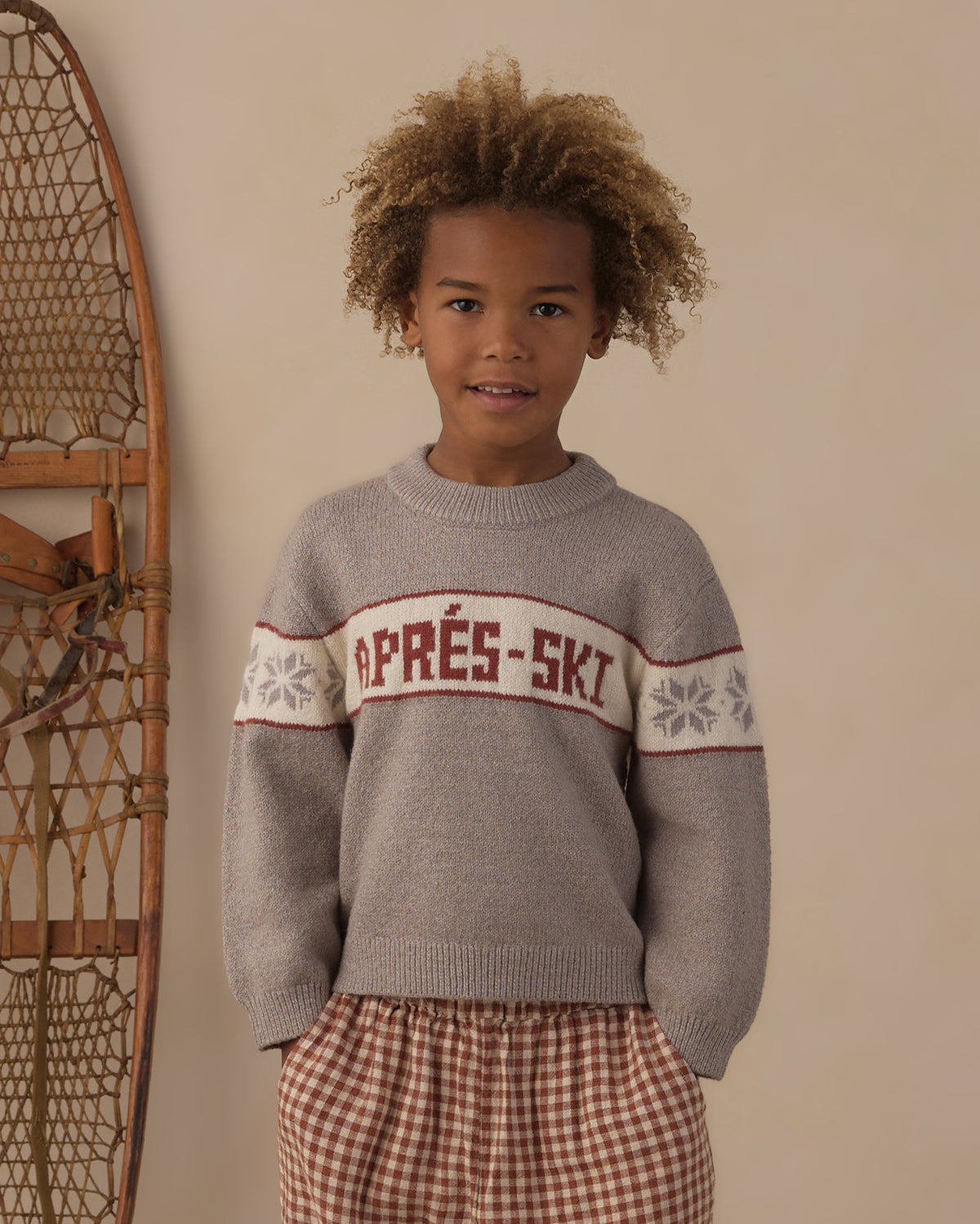 Child in front of vintage snowshoes wearing the Reagan Apres Ski sweater with brick check pants