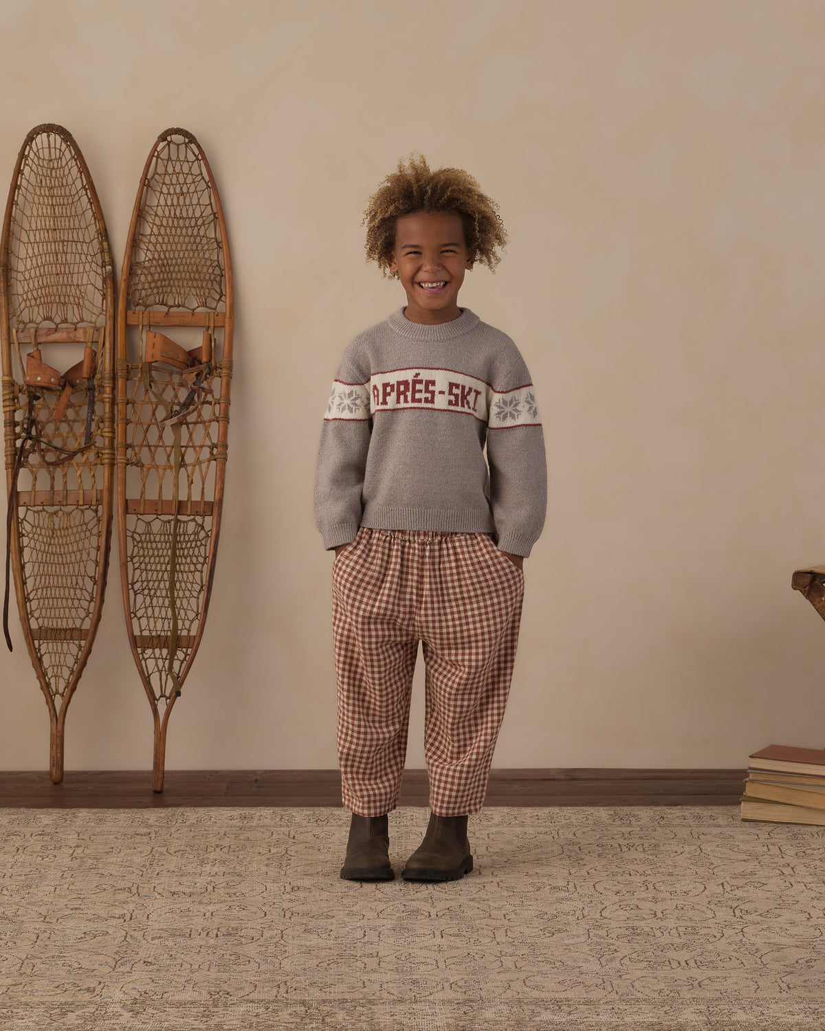 Child standing beside wall-mounted snowshoes in the Reagan Apres Ski sweater and brick check pants