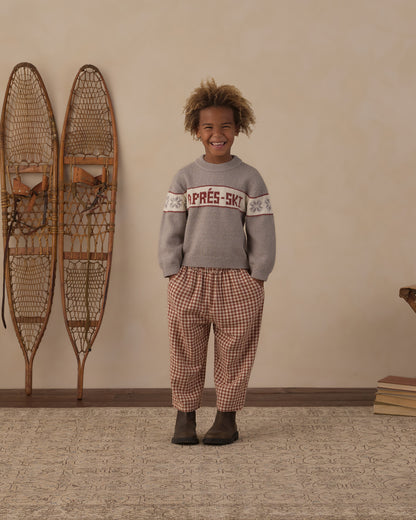 Child standing beside wall-mounted snowshoes in the Reagan Apres Ski sweater and brick check pants
