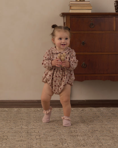 Toddler standing and smiling in the Rylee + Cru Cora Romper in pink floral beside a wooden dresser