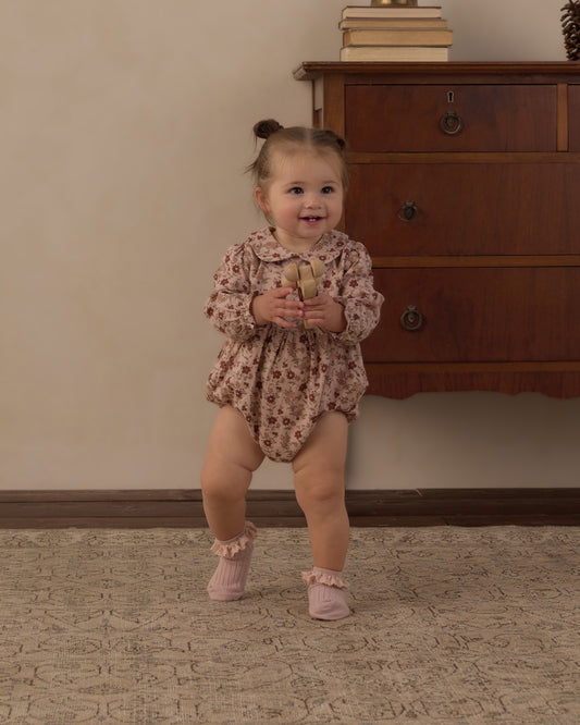 Toddler standing and smiling in the Rylee + Cru Cora Romper in pink floral beside a wooden dresser