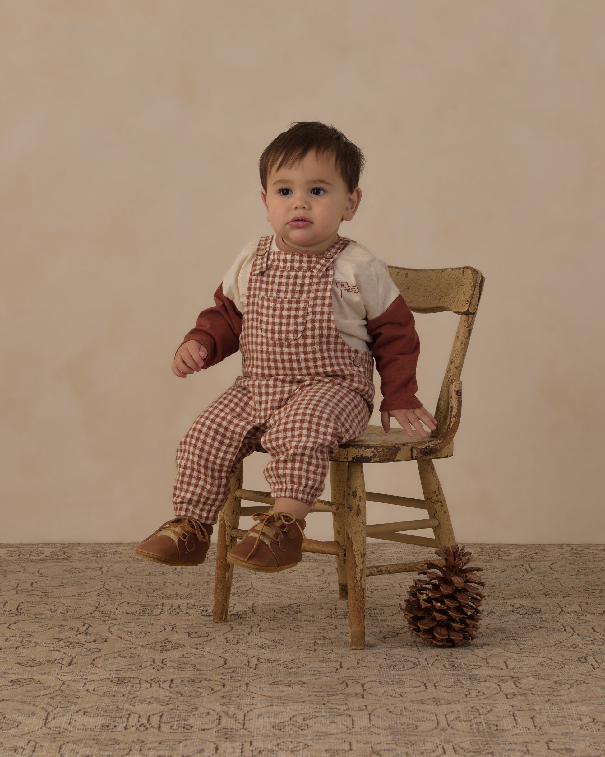 Baby sitting on a wooden chair in the Rylee + Cru Baby Overalls in brick check