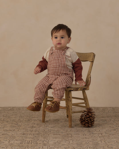 Baby sitting on a wooden chair in the Rylee + Cru Baby Overalls in brick check