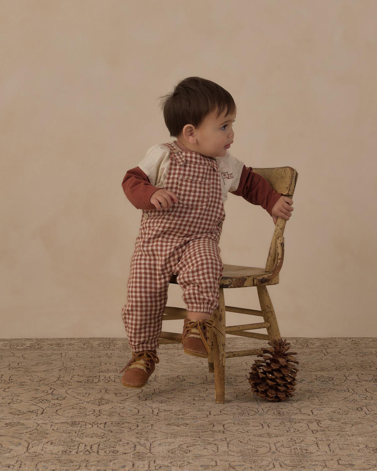 Baby beside a wooden chair wearing the brick check baby overalls and a rust long sleeve top