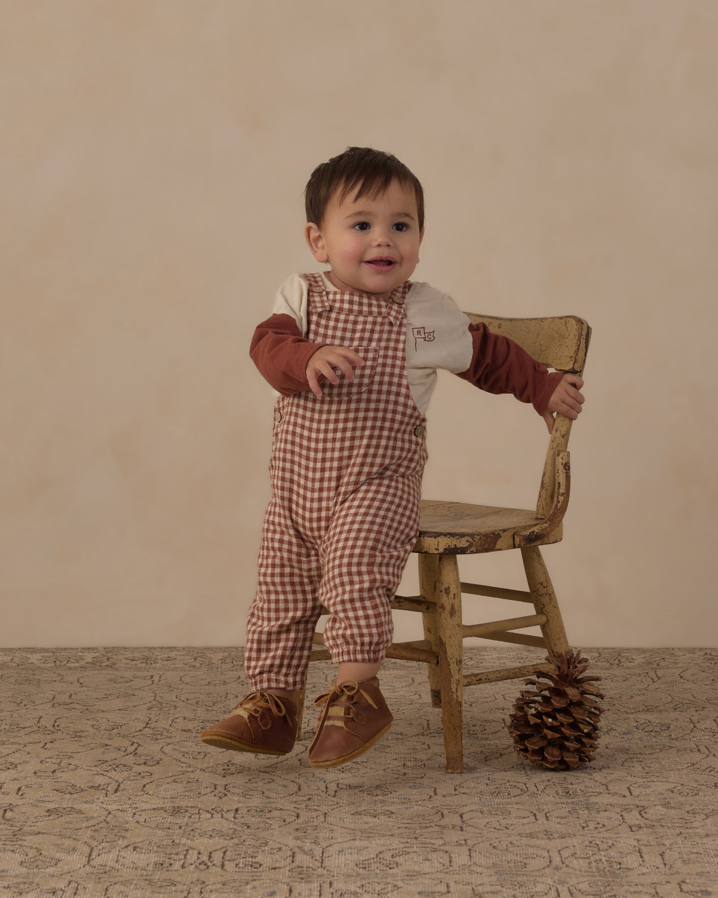 Baby standing and holding a wooden chair in the Rylee + Cru brick check baby overalls