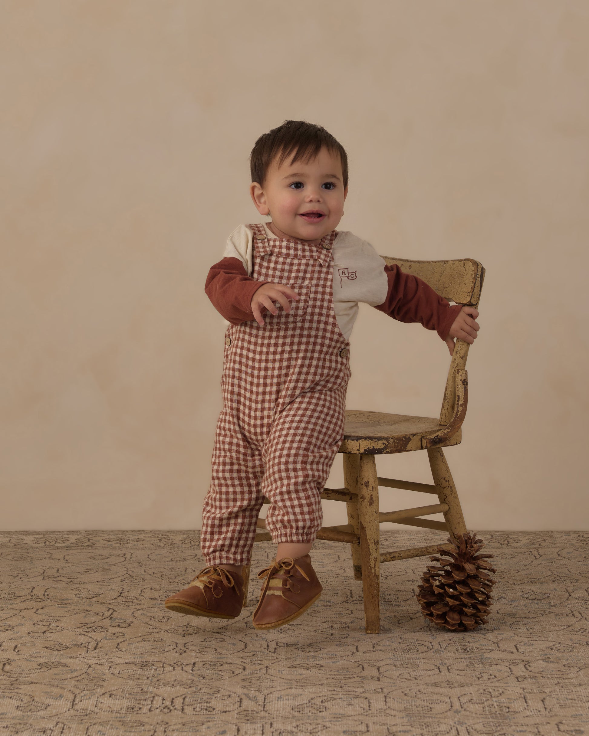 Baby standing and holding a wooden chair in the Rylee + Cru brick check baby overalls