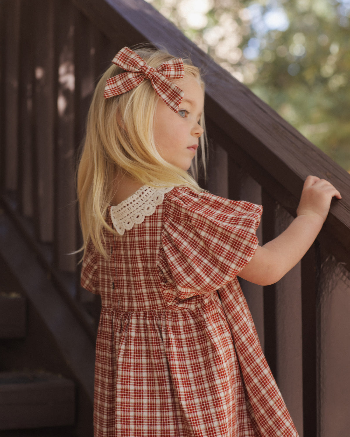 Back view of a girl with a hair bow wearing the Rylee + Cru Heidi dress in vintage plaid on a staircase