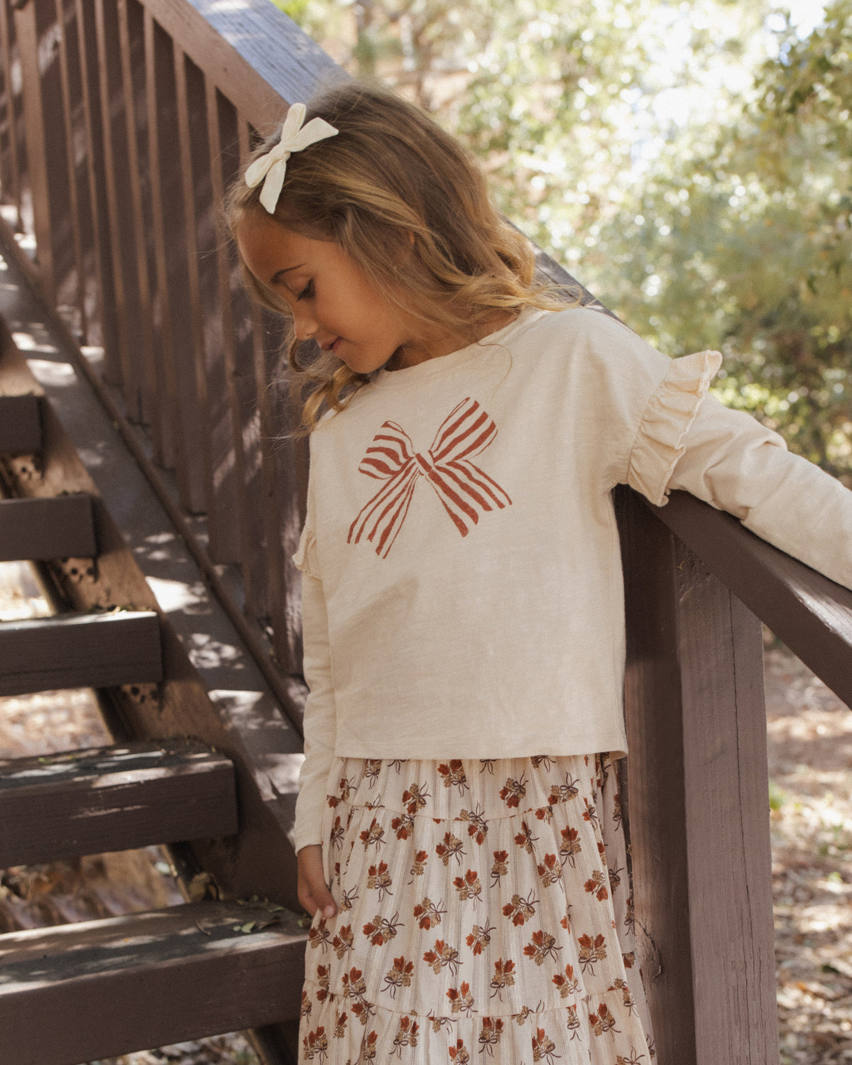 Girl with a hair bow leaning on an outdoor railing in the Rylee + Cru ruffle long sleeve tee with a bow graphic