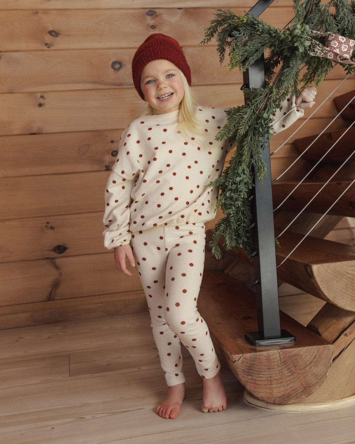 Girl in a red beanie wearing the Rylee + Cru spongey knit set in ruby dots on a wooden staircase with greenery