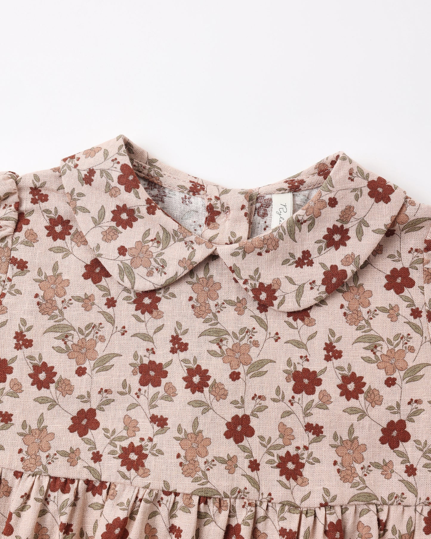 Close-up of a children's romper with a ruffled Peter Pan collar and a dusty floral print on a light beige linen-like fabric, featuring red and brown flowers with green leaves.