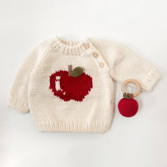 A cream-colored baby sweater with a red apple and green 3D leaf design, featuring three wooden buttons, and a small red apple accessory attached with a wooden ring.