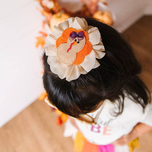 Top of girls head wearing the chiffon Thanksgiving hair clip featuring multicolor turkey motif. The turkey has a small chiffon hair bow on her head. Ribbon-covered alligator clip to grip the hair. 