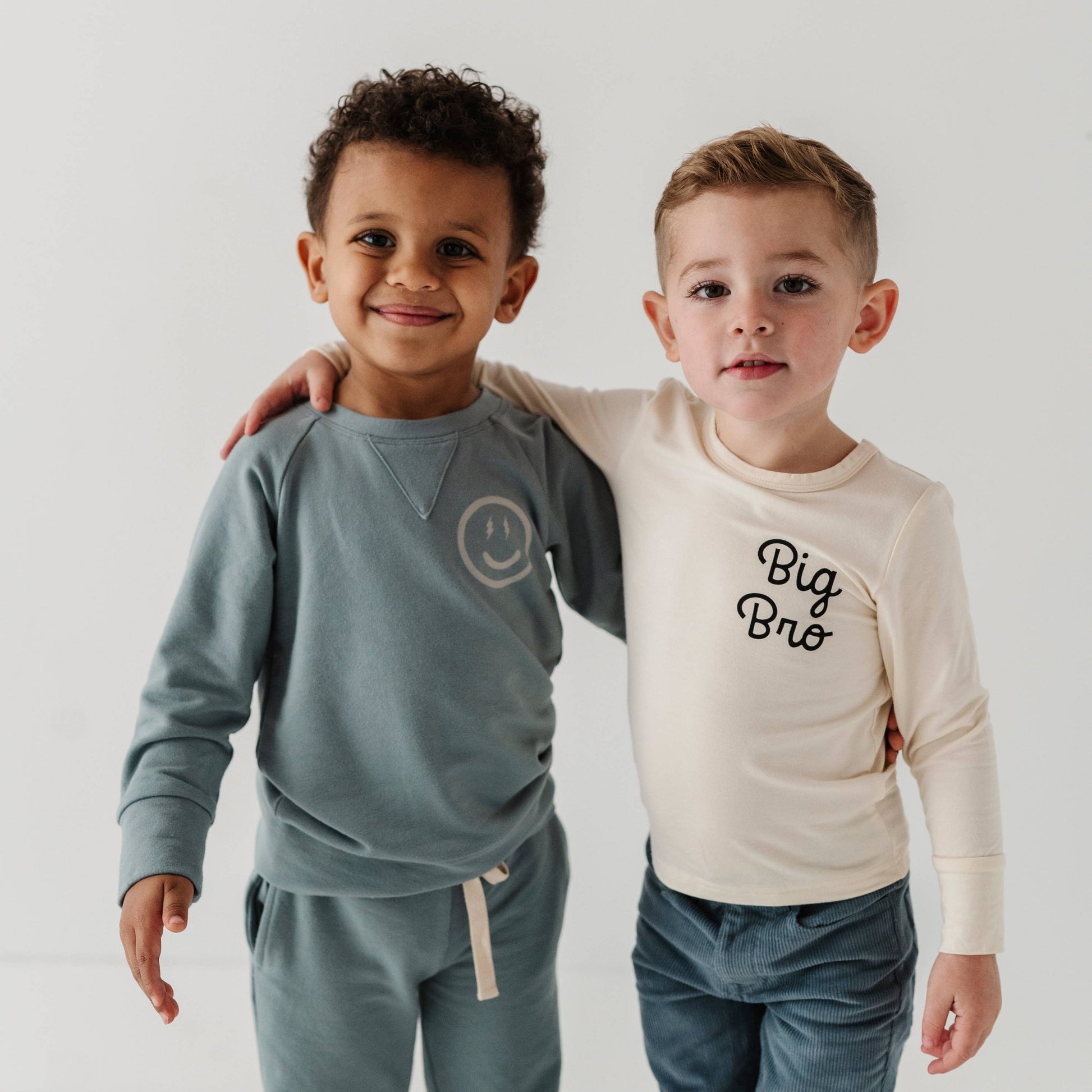 Two boys smiling. Boy on left is wearing our Smile Raglan Sweatshirt and boy on right is wearing our Big Bro longsleeve bamboo tee. 