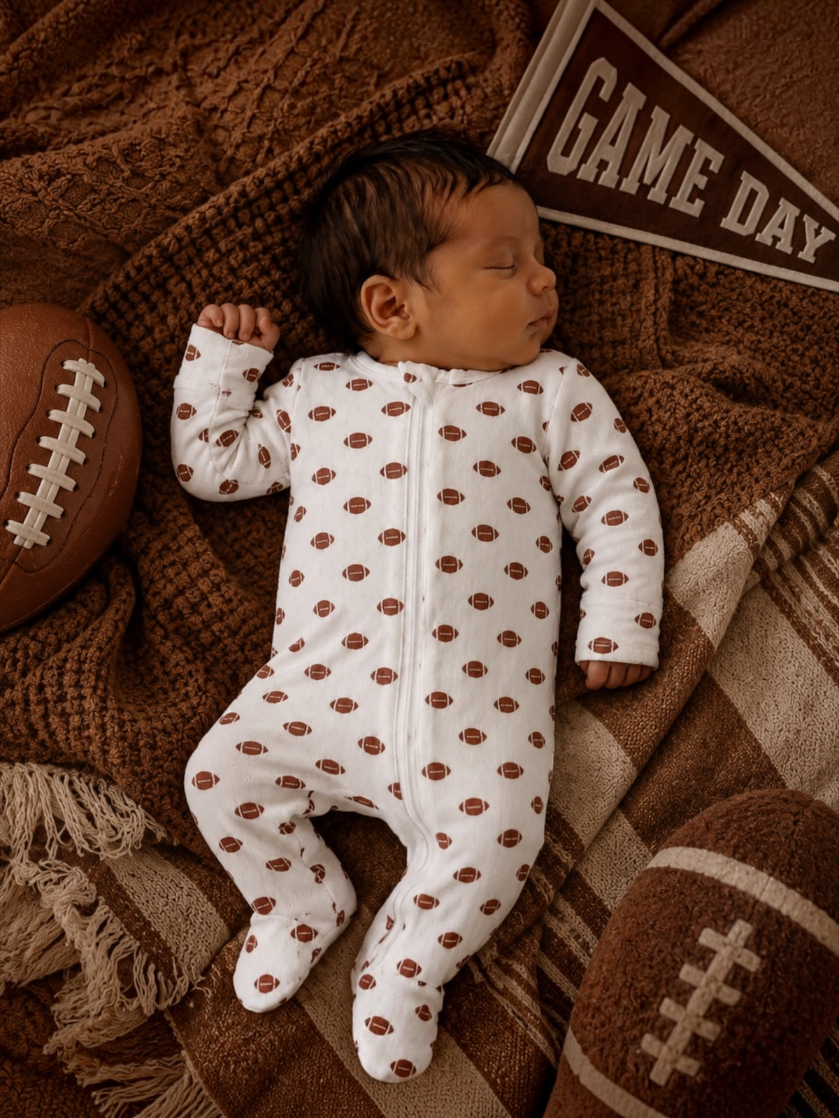 Baby lying on a football wearing the football print organic pointelle two-way zip footie