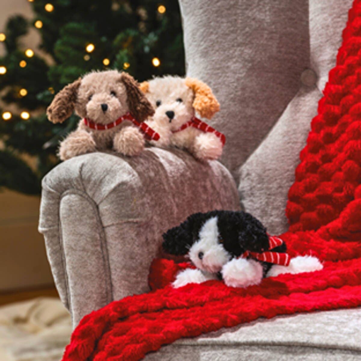 Three small plush dog toys wearing red striped scarves are arranged on a gray upholstered armchair with a chunky red knit blanket, with a blurred Christmas tree in the background.