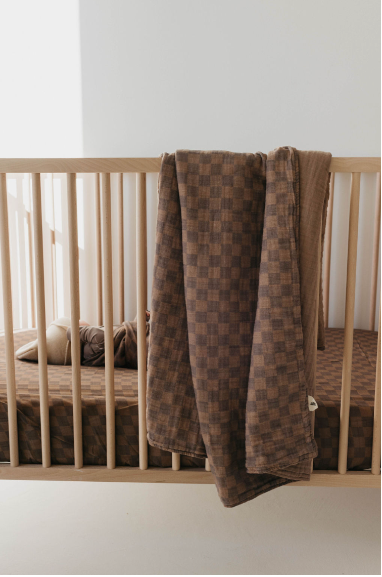 Forever French Baby faded brown checkerboard muslin quilt draped over a natural wood crib