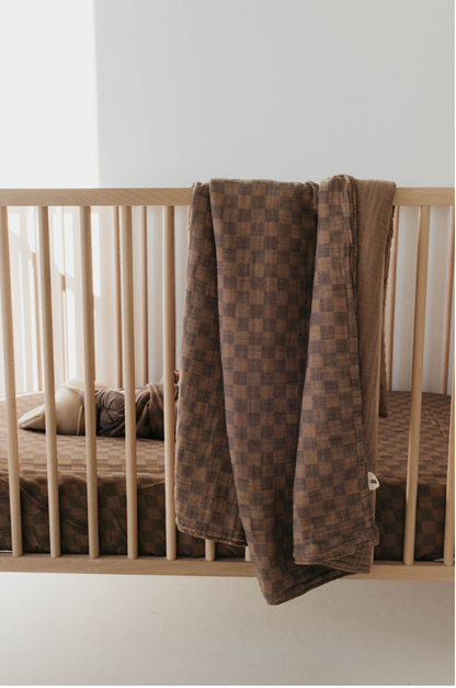Forever French Baby faded brown checkerboard muslin quilt draped over a natural wood crib