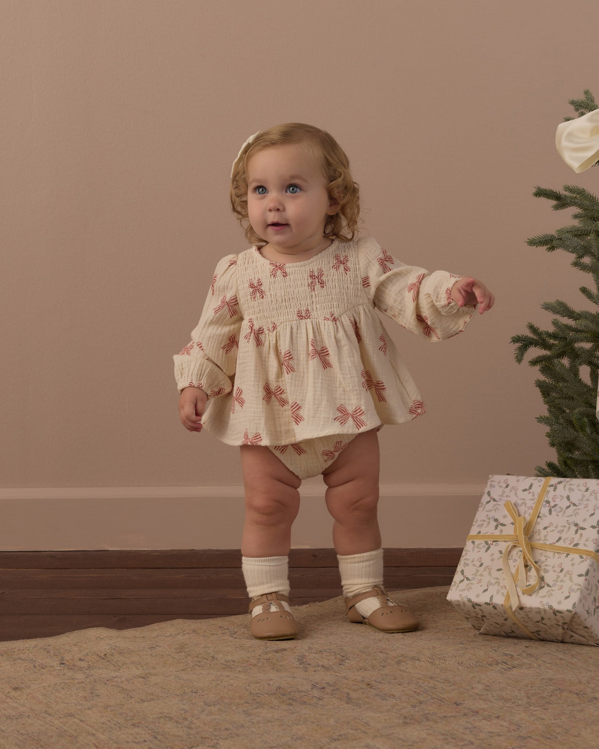 Toddler standing in the Rylee + Cru Sophia top and bloomer set in peppermint bows beside a small Christmas tree and wrapped gift