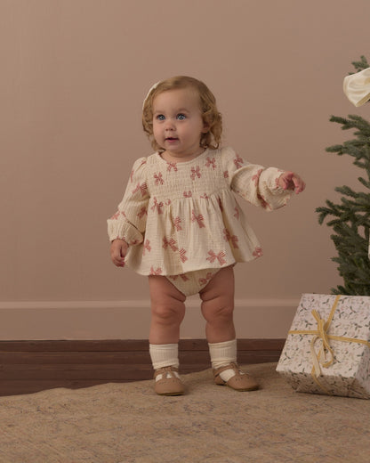Toddler standing in the Rylee + Cru Sophia top and bloomer set in peppermint bows beside a small Christmas tree and wrapped gift