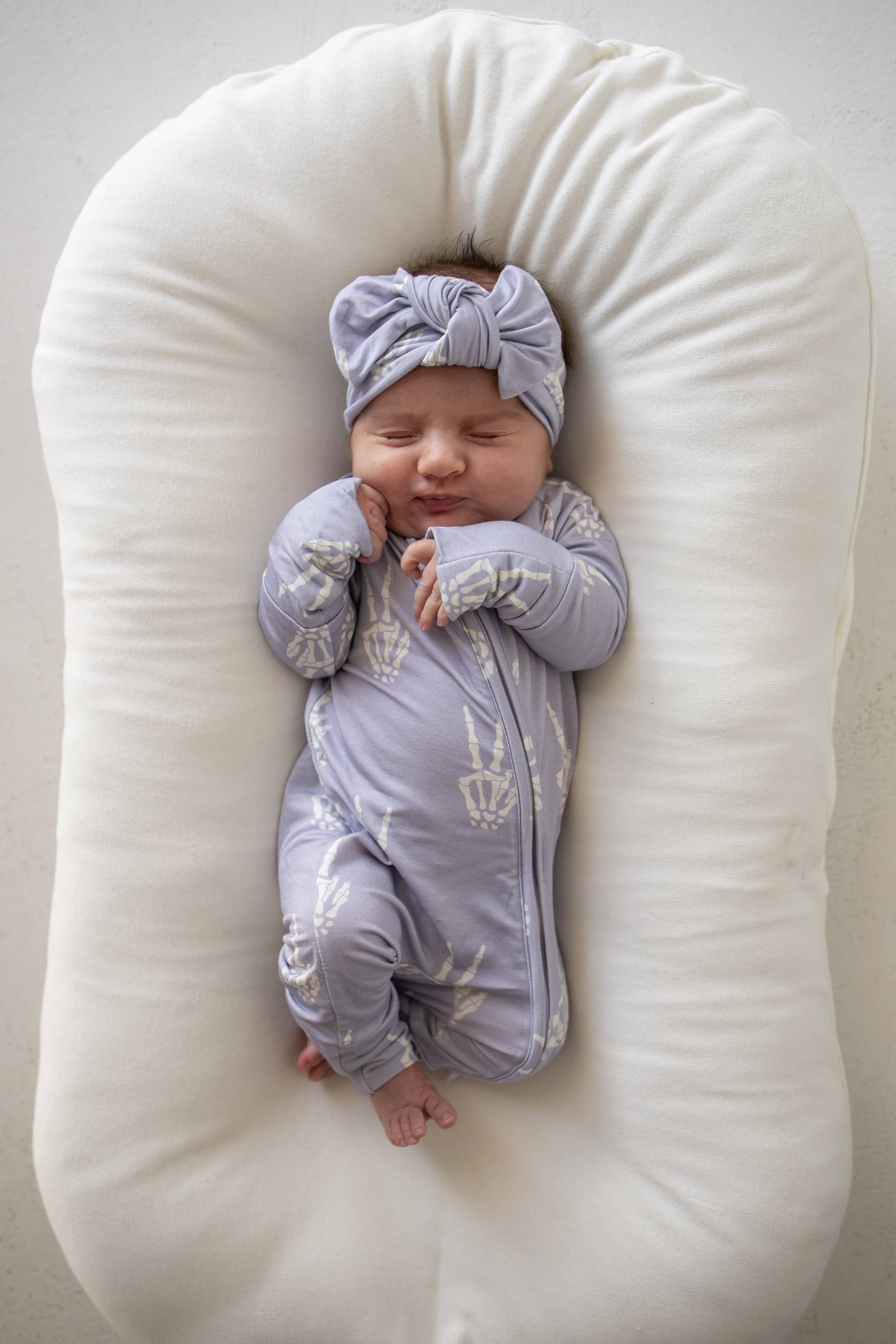 Smiling baby in Enchanted Skelly bamboo zip pajamas and matching bow headband in a lounger