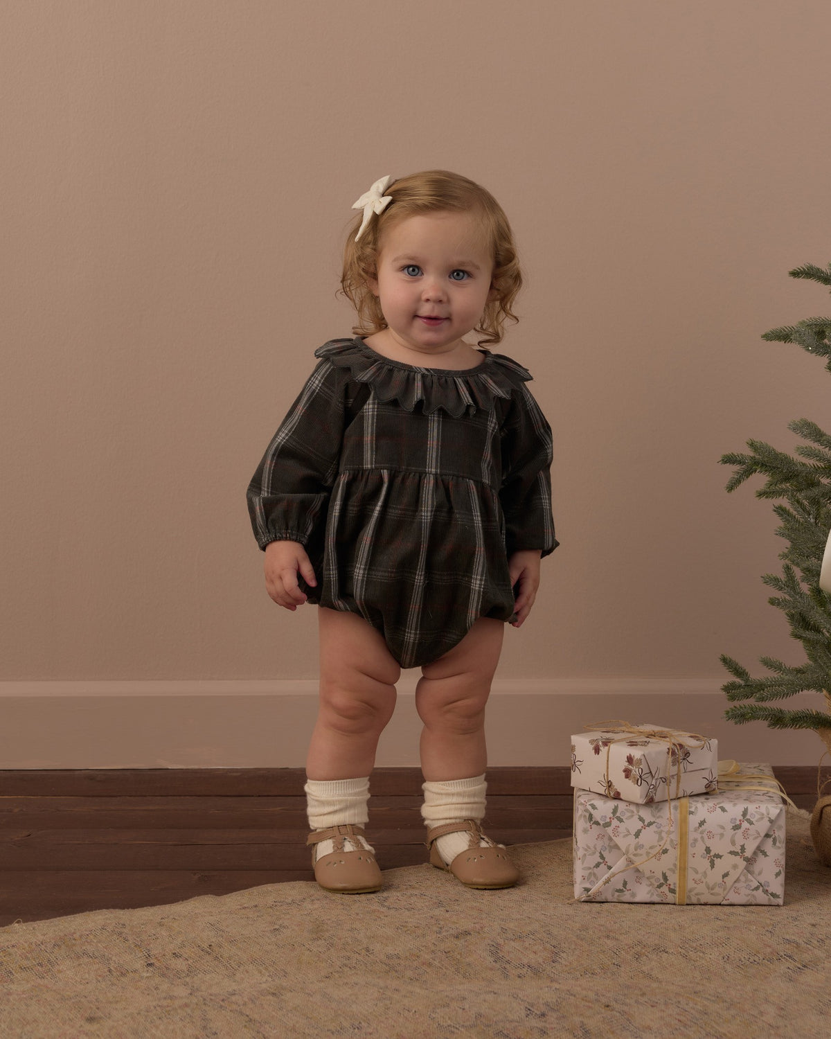 Toddler with a hair bow in the Rylee + Cru Freya romper in dark holiday plaid, front view with a wrapped present at her feet