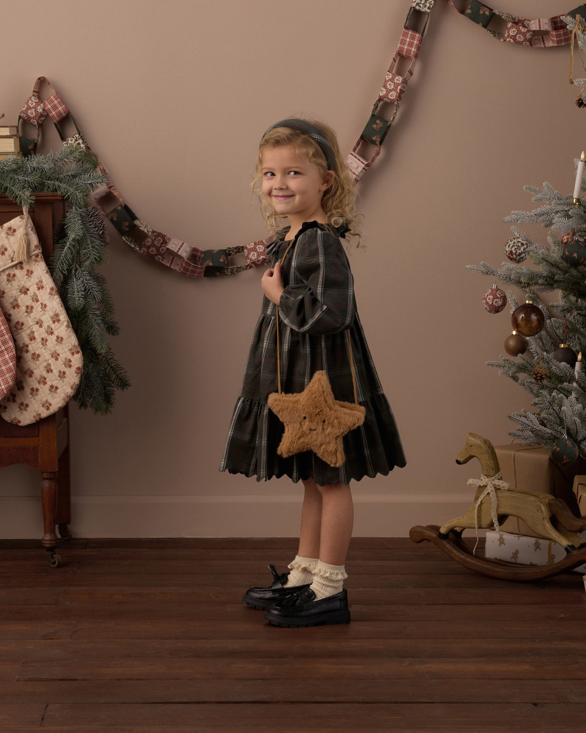 Girl carrying the Rylee + Cru gold star purse over her shoulder while wearing a dark plaid dress in a holiday-decorated room