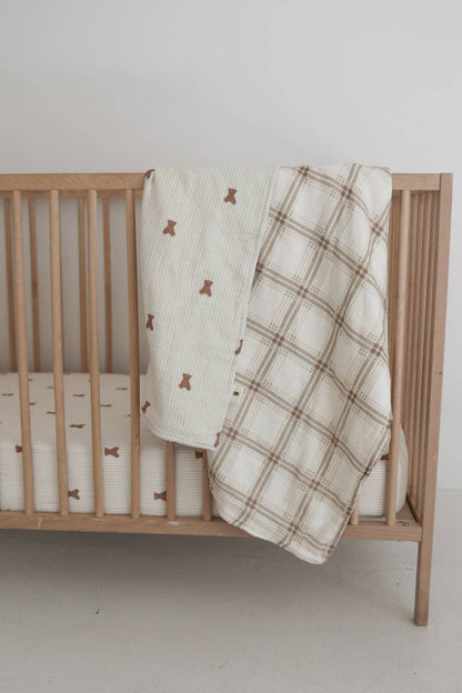 Ivory muslin baby quilt with brown teddy bear print hanging over a natural wood crib