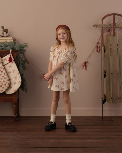 Girl standing in a holiday-decorated room wearing the Rylee + Cru Sabrina mini dress in peppermint bows with a sled behind her