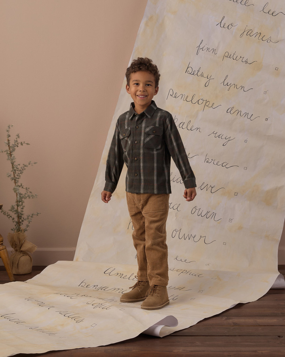 Boy standing on a handwritten backdrop wearing the Rylee + Cru pocketed flannel shirt in holiday plaid with tan pants