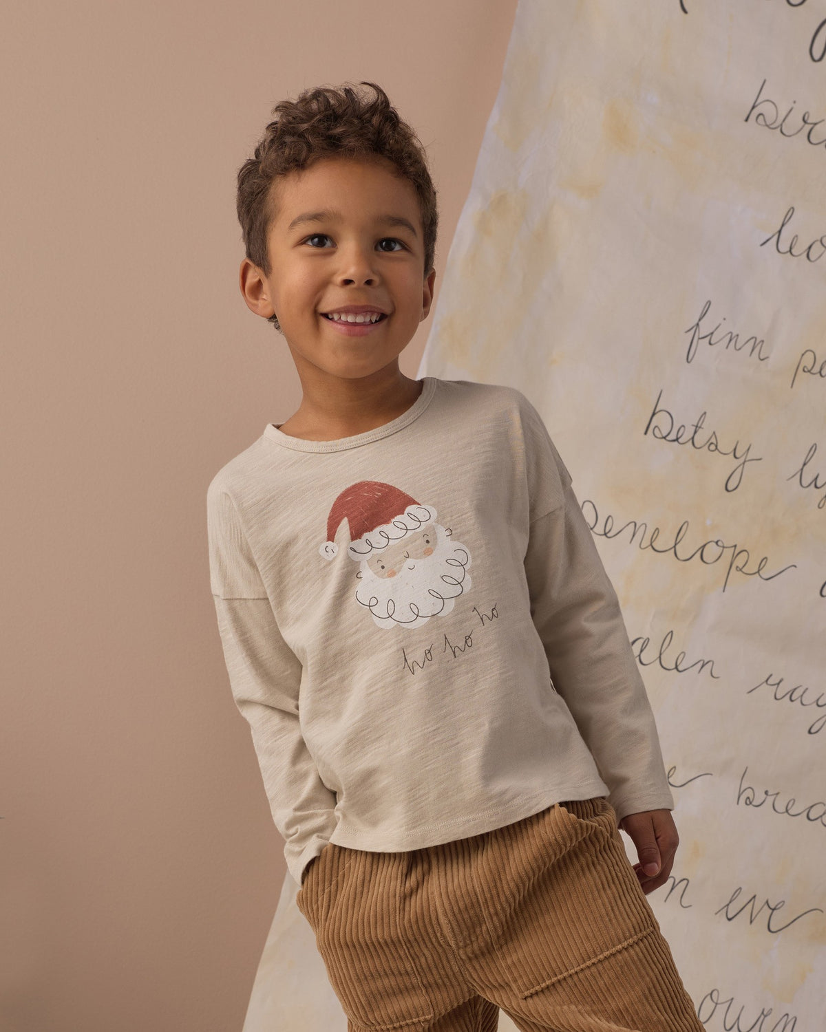 Boy smiling in the Rylee + Cru Santa long sleeve tee with brown corduroy pants in front of a handwritten backdrop