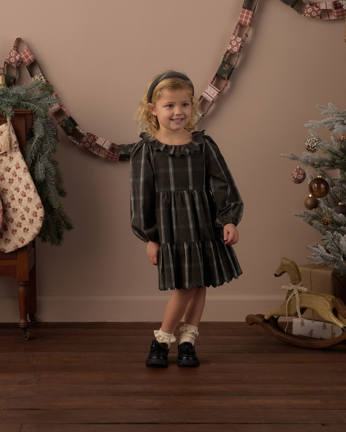 Girl standing under a holiday garland wearing the Rylee + Cru Fran dress in holiday plaid with a rocking horse behind her