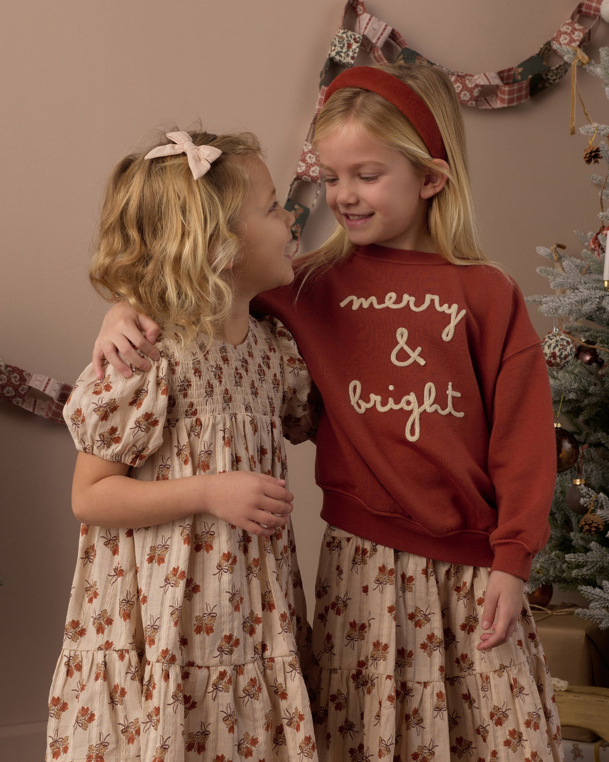 Two girls in holiday outfits, one in the Rylee + Cru Merry & Bright relaxed sweatshirt, standing in front of a Christmas tree