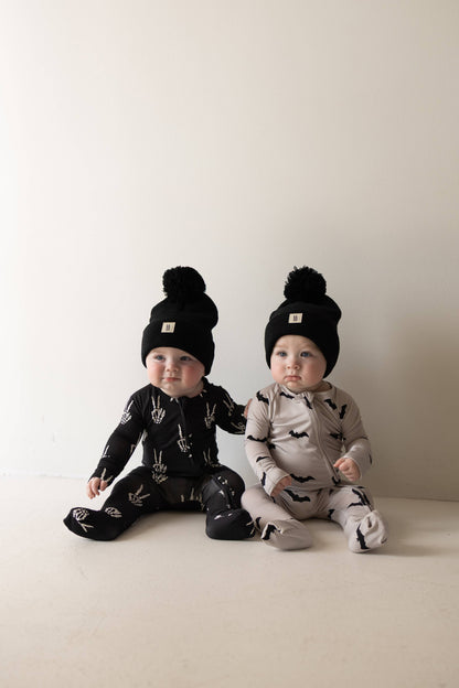 Two babies in Skelly bamboo pajamas and pom beanies, one in Midnight black and one in a lighter colorway