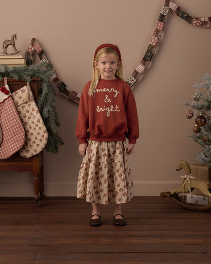 Girl wearing the Rylee + Cru Merry & Bright relaxed sweatshirt over a floral skirt beside a decorated Christmas tree