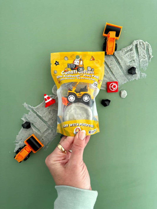 Hand holding the KidDoughs Construction Mini Play Pack pouch with gray dough and toy excavators laid out on green