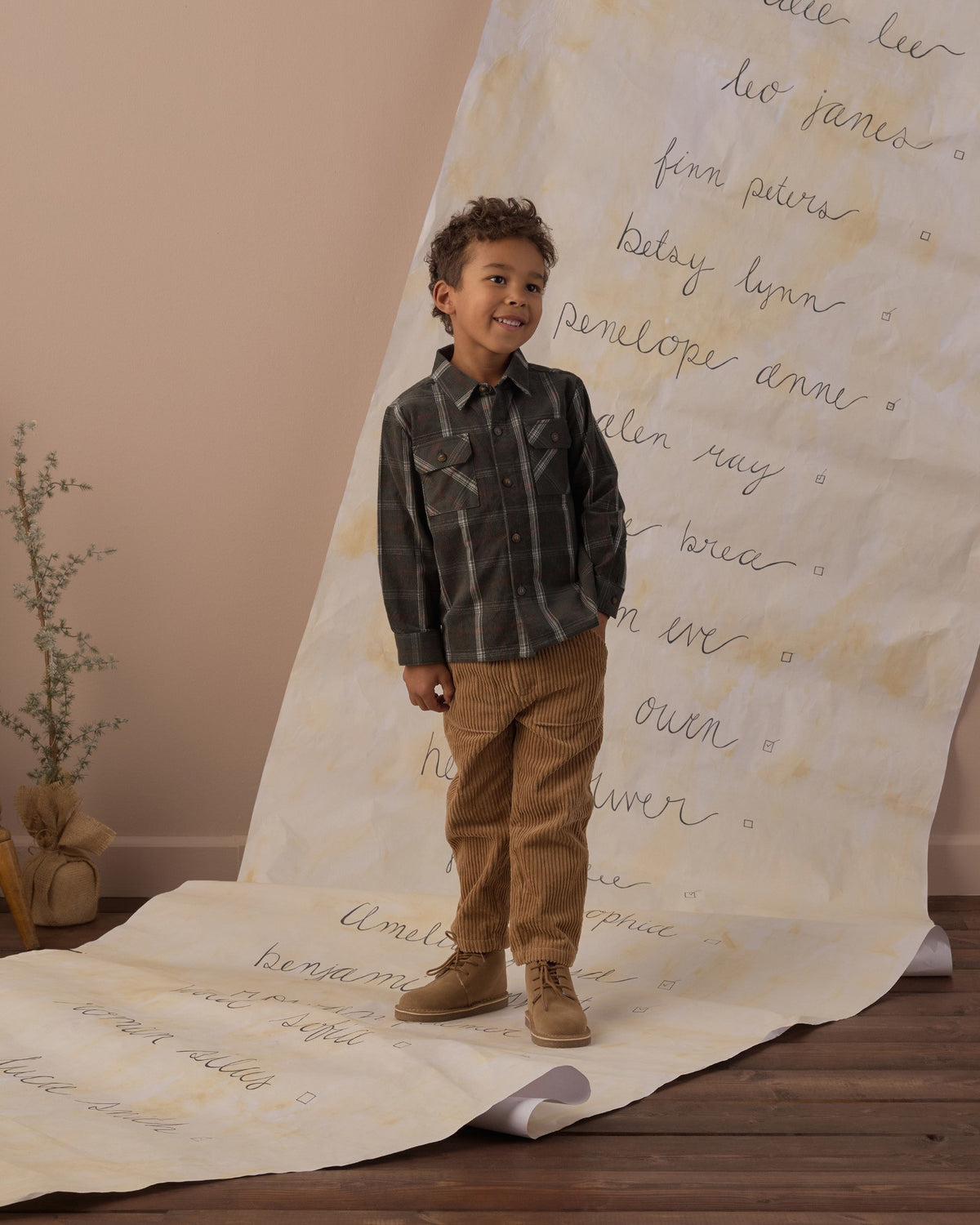 Full-length view of a boy in the Rylee + Cru pocketed flannel shirt in holiday plaid beside a small Christmas tree