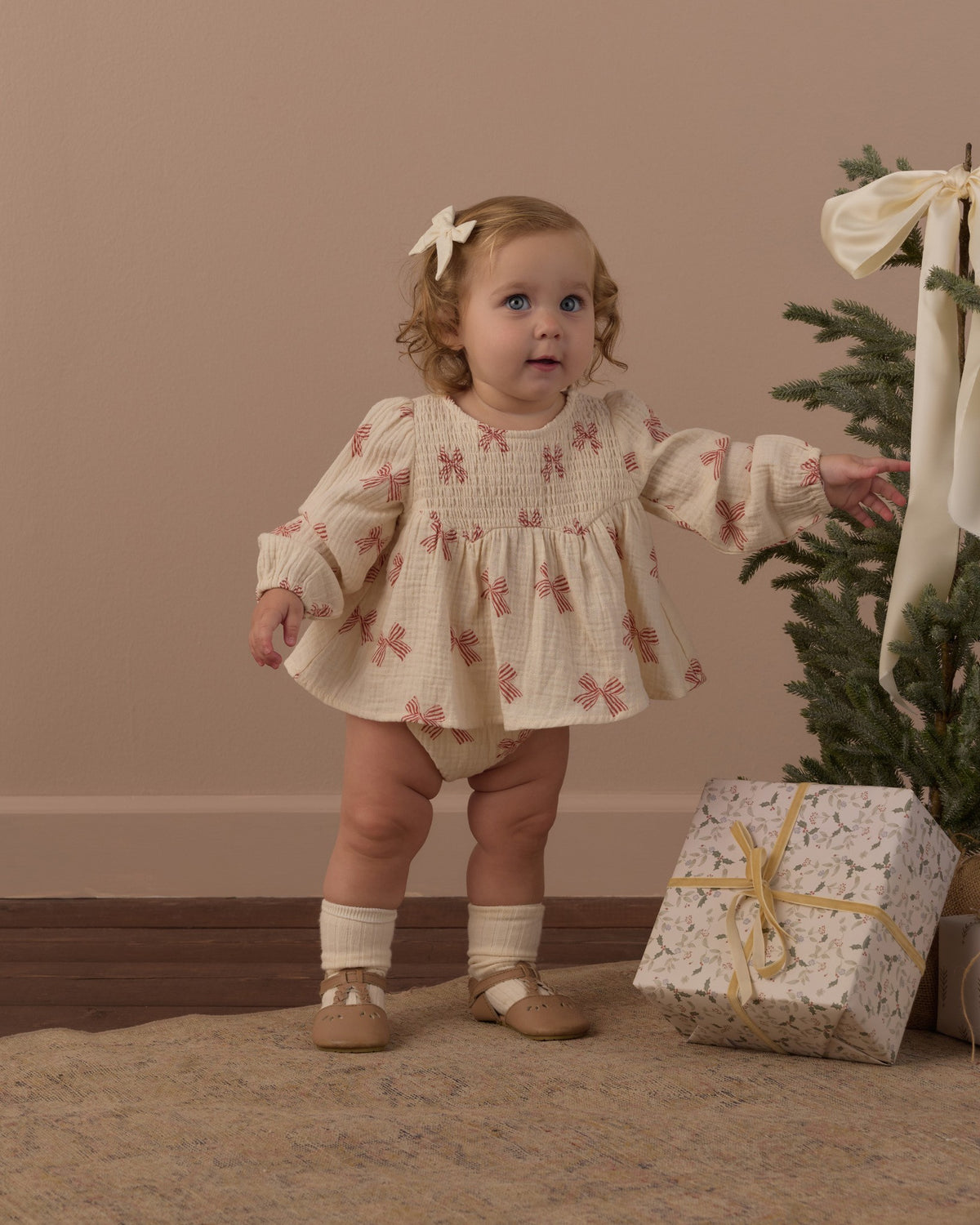 Toddler mid-step in the Rylee + Cru Sophia top and bloomer set in peppermint bows with a wrapped present and Christmas tree