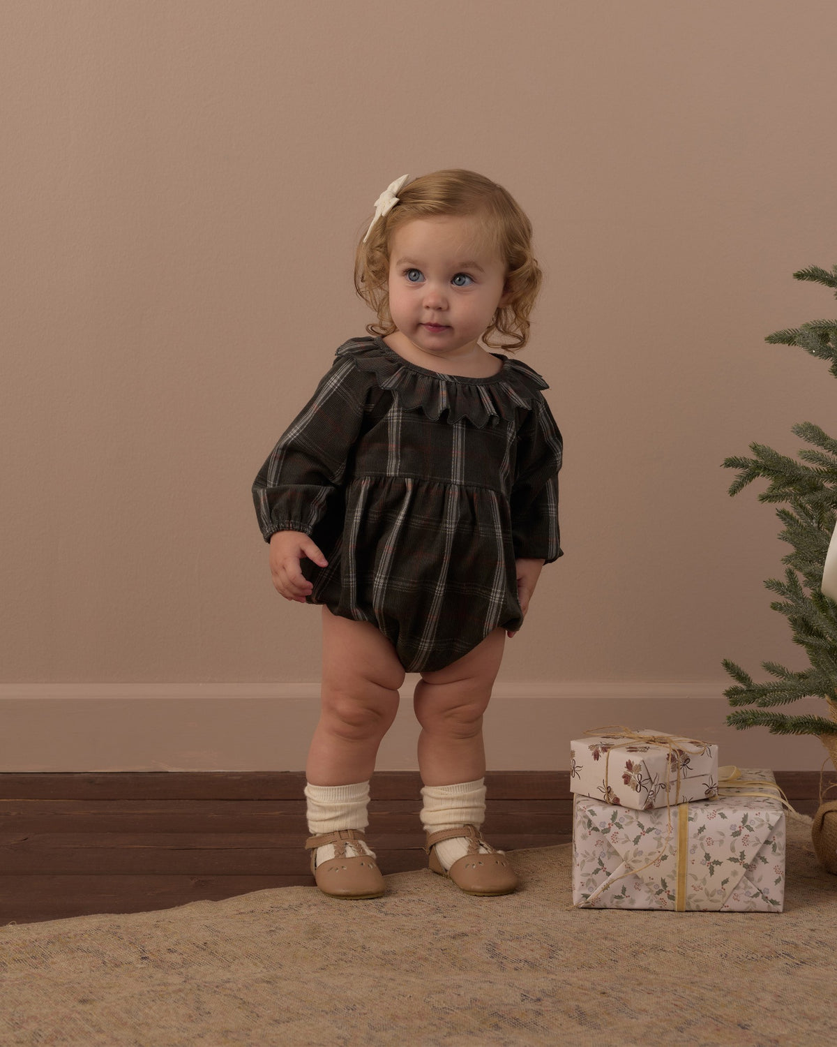 Toddler standing in the Rylee + Cru Freya romper in holiday plaid with socks and shoes beside a small Christmas tree and gift