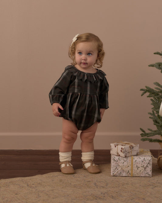 Toddler standing in the Rylee + Cru Freya romper in holiday plaid with socks and shoes beside a small Christmas tree and gift